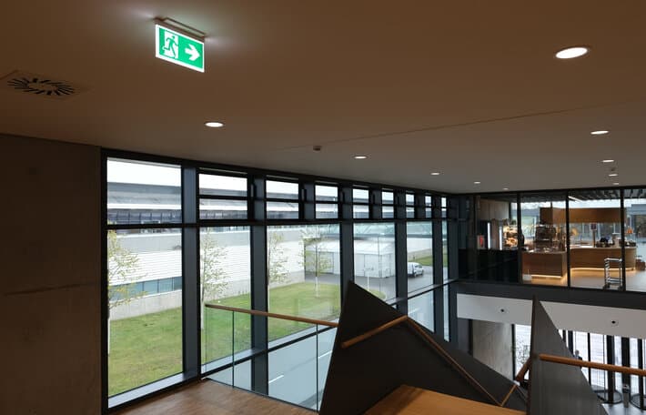 Illuminated emergency exit sign above a mezzanine walkway in a modern commercial building
